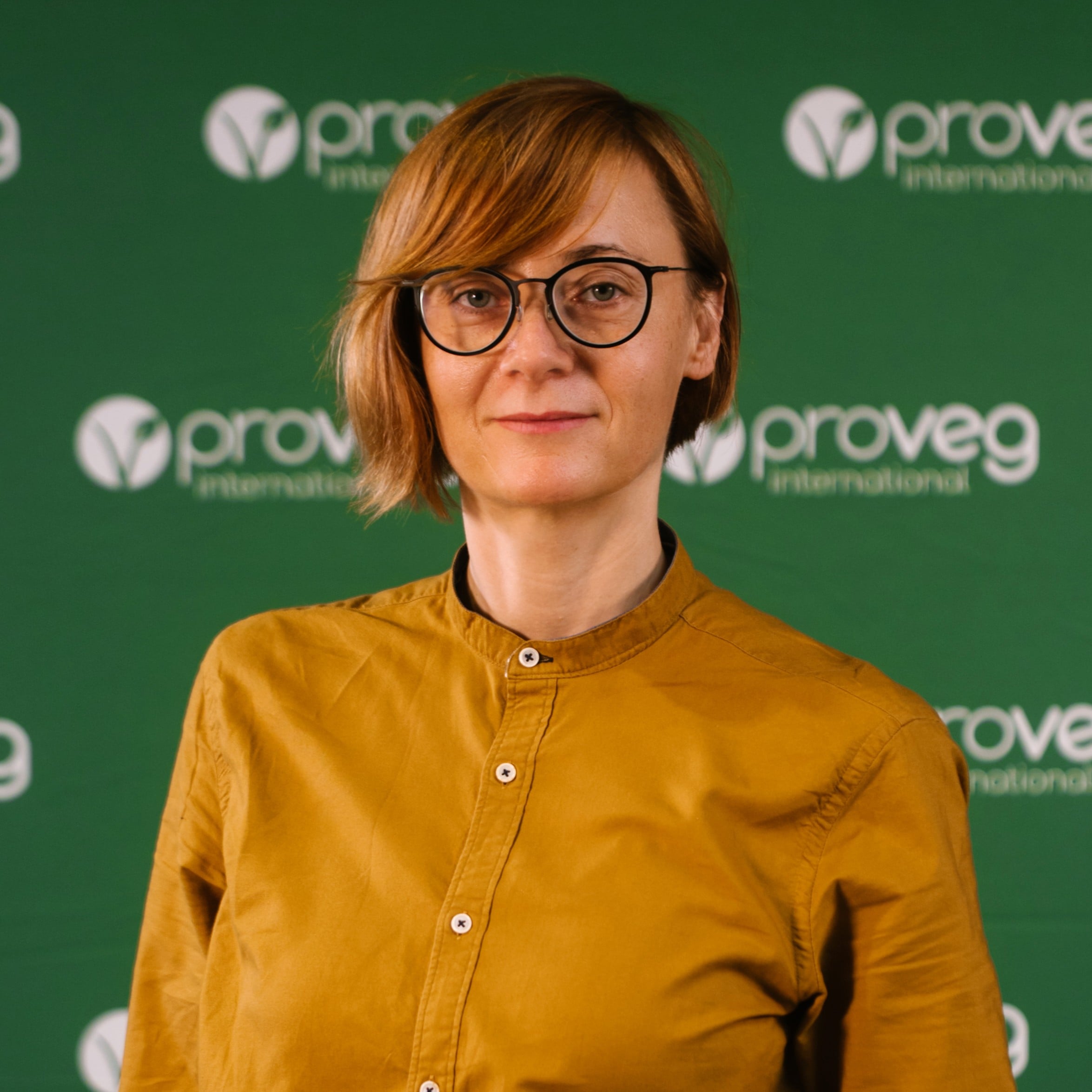 Przed zielonym tłem z logo ProVeg International stoi osoba o krótkich, jasnobrązowych włosach i w okularach, ubrana w koszulę w kolorze musztardowo-żółtym, reprezentująca Szkołę Przyszłości.