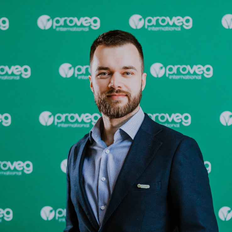Mężczyzna w garniturze stoi przed zielonym tłem z powtarzającymi się logo ProVeg International, symbolizującymi współpracę między ProVeg a Szkołą Przyszłości.