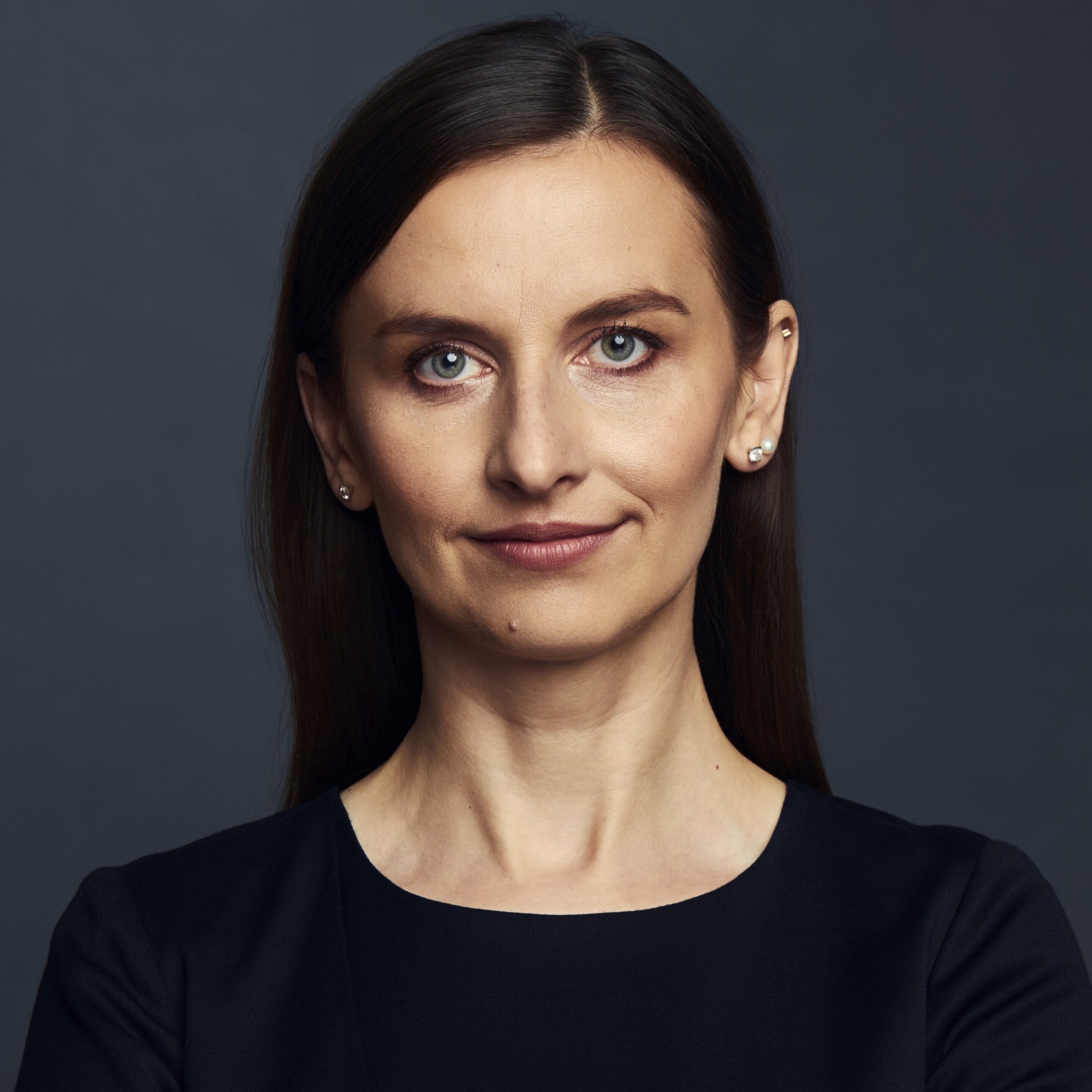 Kobieta o prostych brązowych włosach, ubrana w czarną bluzkę, pozuje na jednolitym ciemnym tle, patrząc prosto w obiektyw z neutralnym wyrazem twarzy — jej opanowana pewność siebie odzwierciedla innowacyjnego ducha Szkoły Przyszłości.