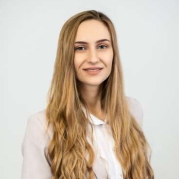 Kobieta o długich, falujących blond włosach uśmiecha się lekko. Ma na sobie białą bluzkę i jasną marynarkę; stoi na prostym, jasnym tle, uosabiając profesjonalnego ducha Szkoły Przyszłości.