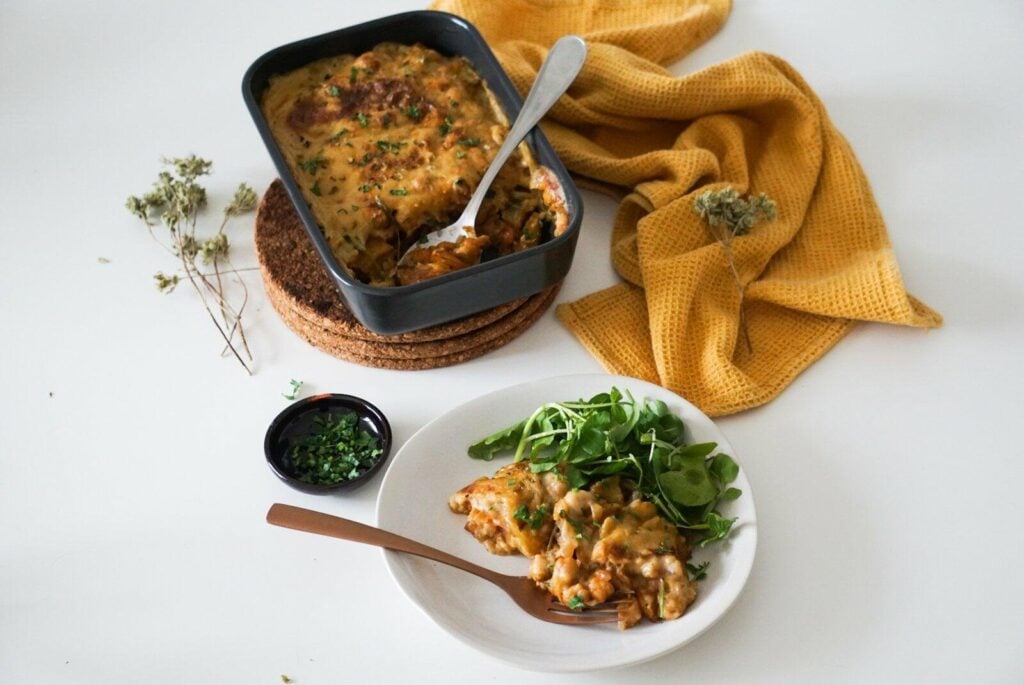 Caçarola assada em um prato preto com uma porção em um prato branco ao lado. O prato é guarnecido com ervas. Uma pequena tigela de verduras picadas e um pano amarelo mostarda também estão presentes em uma superfície branca.