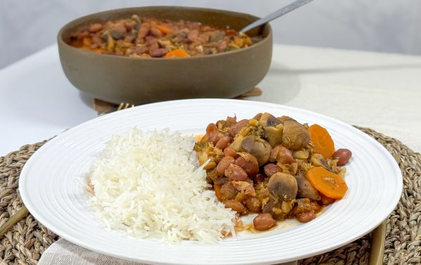 Um prato branco com arroz cozido no vapor e um guisado de legumes e feijão - feijão, cenoura, cogumelos e molho - servido em um jogo americano de tecido; ao fundo, um pote com esta receita vegetariana portuguesa.
