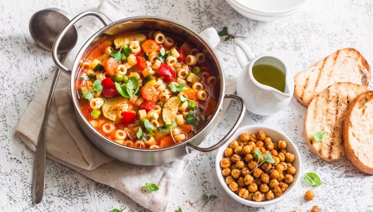 Uma panela de sopa de macarrão com legumes e ervas, servida com pão grelhado, uma tigela de grão-de-bico assado e uma pequena jarra de azeite de oliva em uma superfície texturizada.