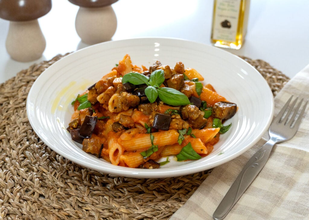Uma tigela branca de macarrão penne com molho de tomate, berinjela assada e manjericão fresco, colocada sobre uma esteira trançada, com um garfo, um guardanapo e uma garrafa de azeite de oliva ao lado.