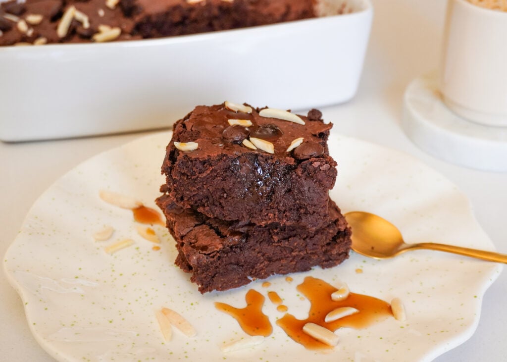 Dois brownies de chocolate empilhados em um prato branco, decorados com lascas de amêndoa e calda de caramelo, com uma colher dourada ao lado deles. Uma assadeira branca aparece parcialmente no fundo.