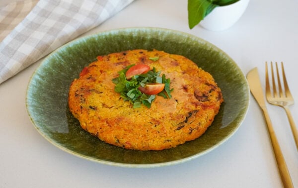 Uma panqueca redonda de vegetais, decorada com tomates-cereja e ervas, servida em um prato verde.