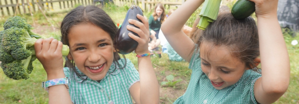 Two young girls in green checkered uniforms smile and hold vegetables—broccoli, eggplant, and zucchini—above their heads while playing outdoors in a garden, with other children blurred in the background.