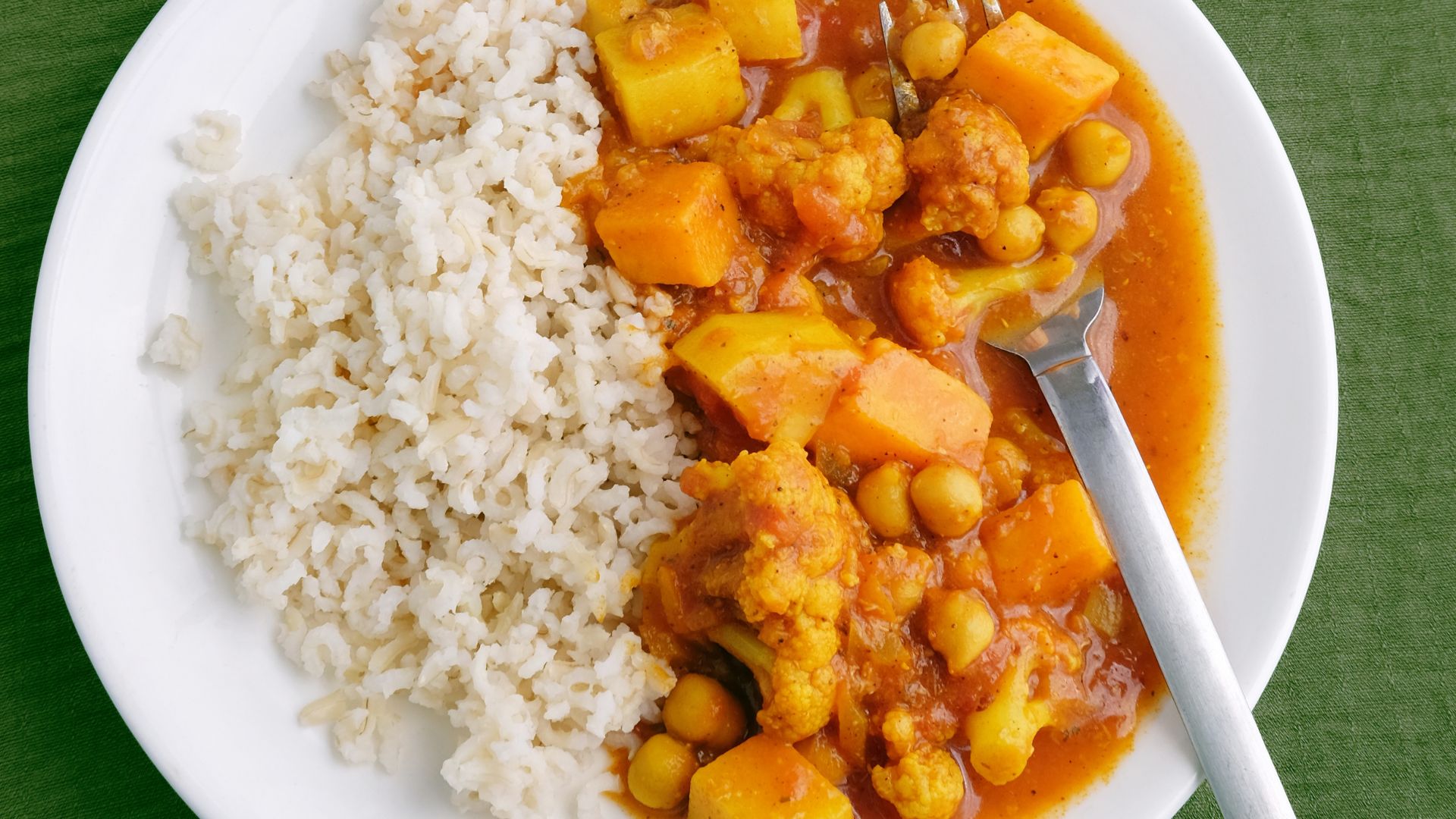 A plate with brown rice on one side and vegetable curry with chickpeas, cauliflower, and potatoes in a tomato-based sauce on the other, with a fork.