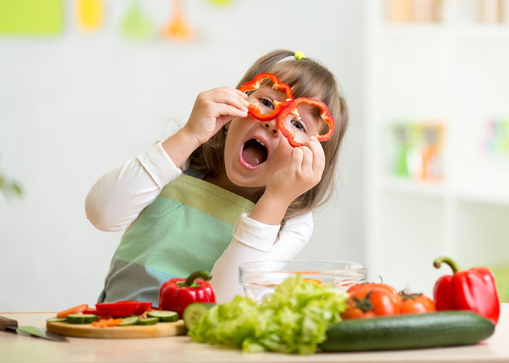 A young child holds red pepper slices over their eyes whilst sitting at a table with various fresh vegetables.