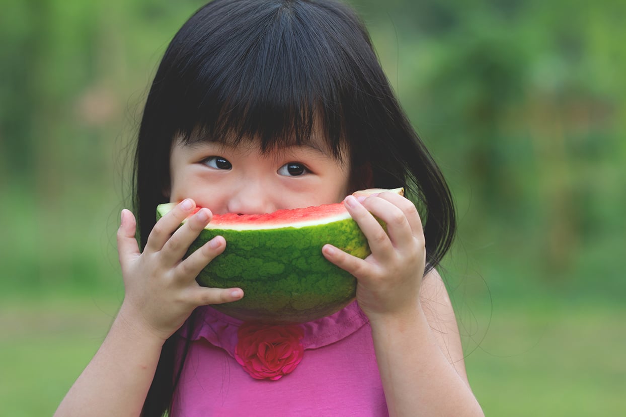 Young girl in a pink dress holding and eating a slice of watermelon outdoors, with green blurred background.