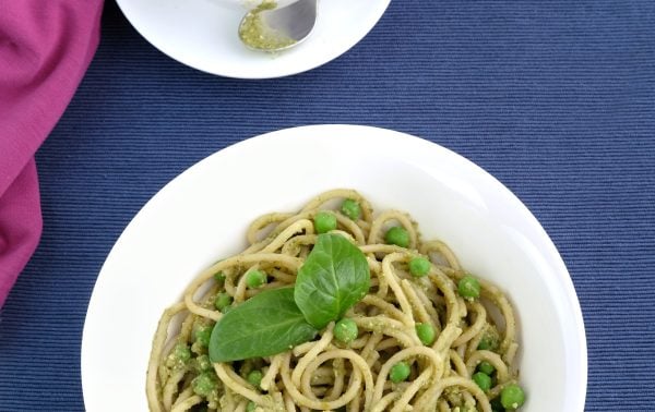 A white bowl of spaghetti with green peas and pesto, garnished with basil, sits on a blue surface beside a small bowl of pesto with a spoon. A pink cloth is partially visible.