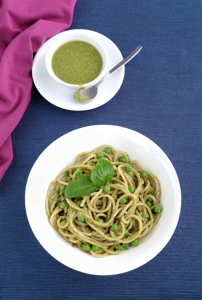 A white bowl of spaghetti with green peas and pesto, garnished with basil, sits on a blue surface beside a small bowl of pesto with a spoon. A pink cloth is partially visible.