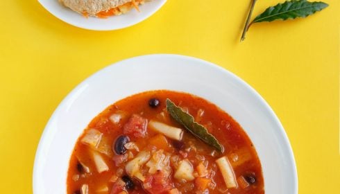 A bowl of vegetable and bean soup garnished with a bay leaf, served with a plate of bread and cheese; two bay leaves are placed on the yellow background.