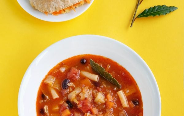 A bowl of vegetable and bean soup garnished with a bay leaf, served with a plate of bread and cheese; two bay leaves are placed on the yellow background.