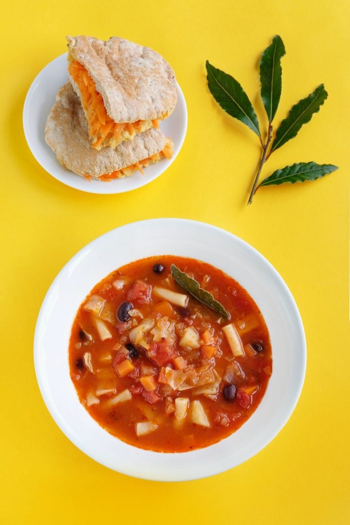A bowl of vegetable and bean soup garnished with a bay leaf, served with a plate of bread and cheese; two bay leaves are placed on the yellow background.