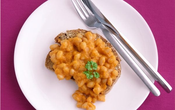 A slice of toast topped with baked beans and a sprig of parsley on a white plate, with a fork and knife, set on a magenta surface.