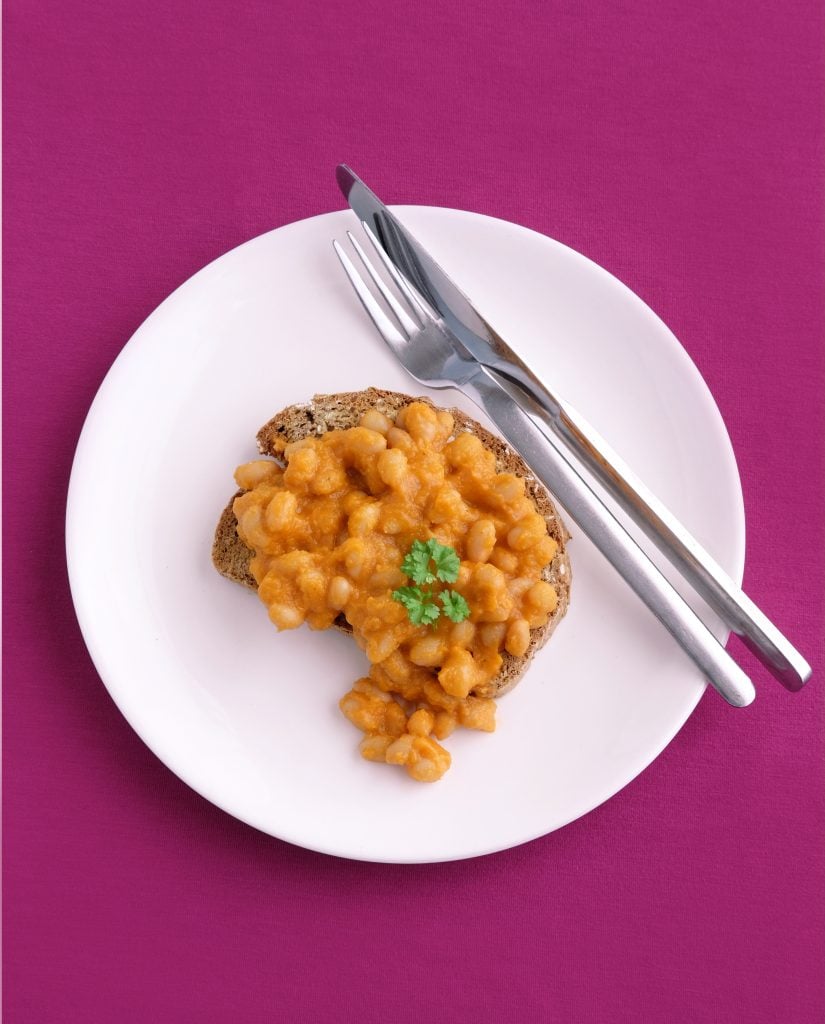 A slice of toast topped with baked beans and a sprig of parsley on a white plate, with a fork and knife, set on a magenta surface.