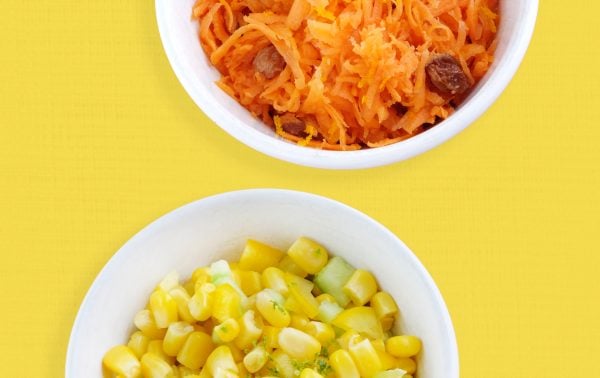 Two white bowls on a yellow background: the top bowl contains grated carrots with sultanas; the bottom bowl contains sweetcorn mixed with small pieces of cucumber.