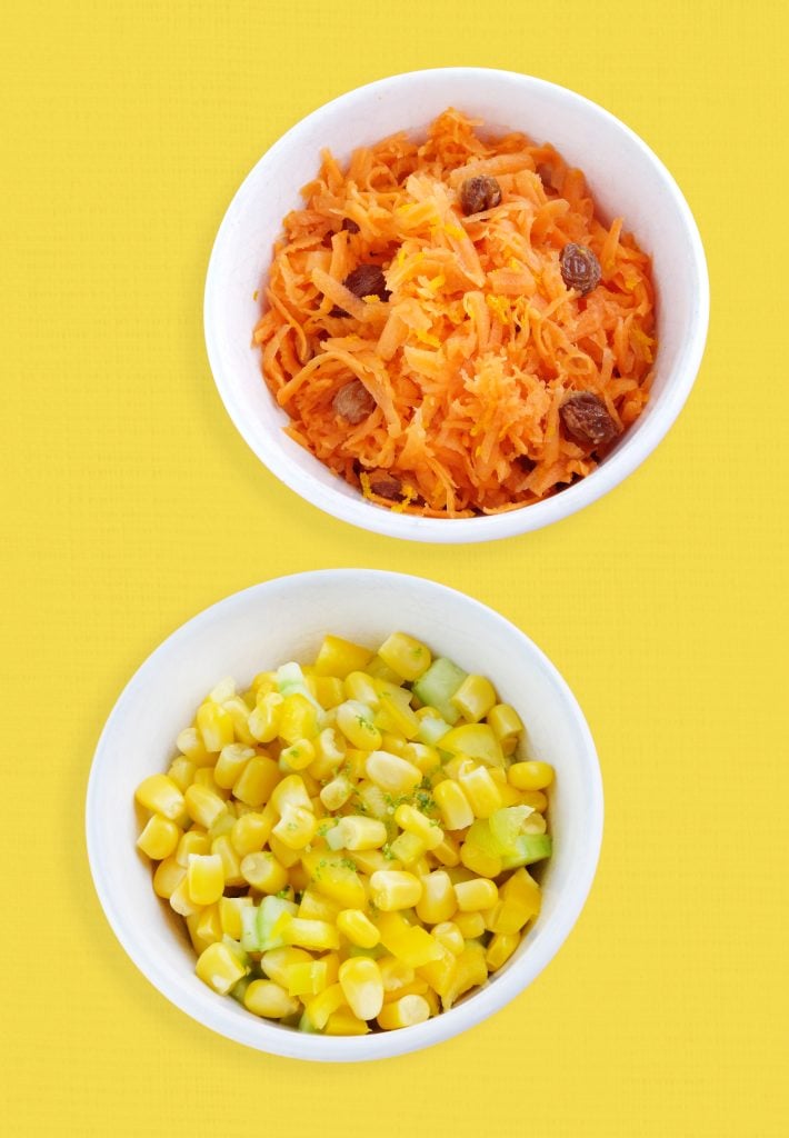 Two white bowls on a yellow background: the top bowl contains grated carrots with sultanas; the bottom bowl contains sweetcorn mixed with small pieces of cucumber.
