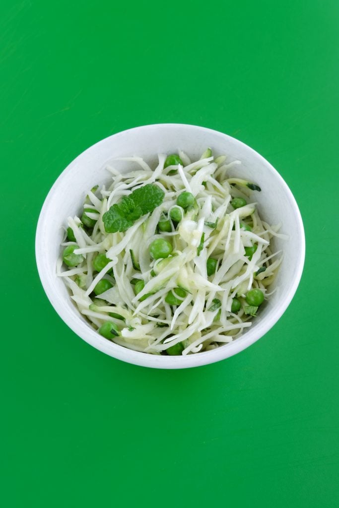 A white bowl filled with shredded cabbage, garden peas, and mint leaves on a solid green background.