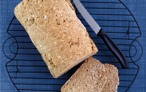 A loaf of wholegrain bread with two slices cut, placed on a round cooling rack beside a bread knife, on a blue textured surface.