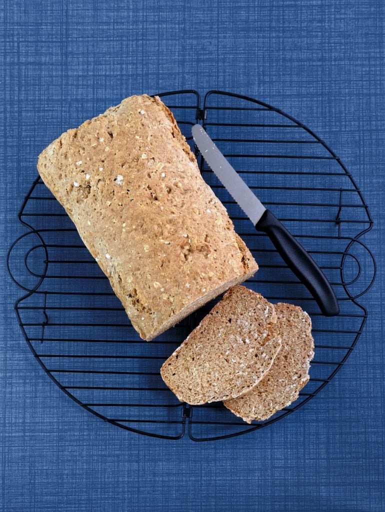 A loaf of wholegrain bread with two slices cut, placed on a round cooling rack beside a bread knife, on a blue textured surface.