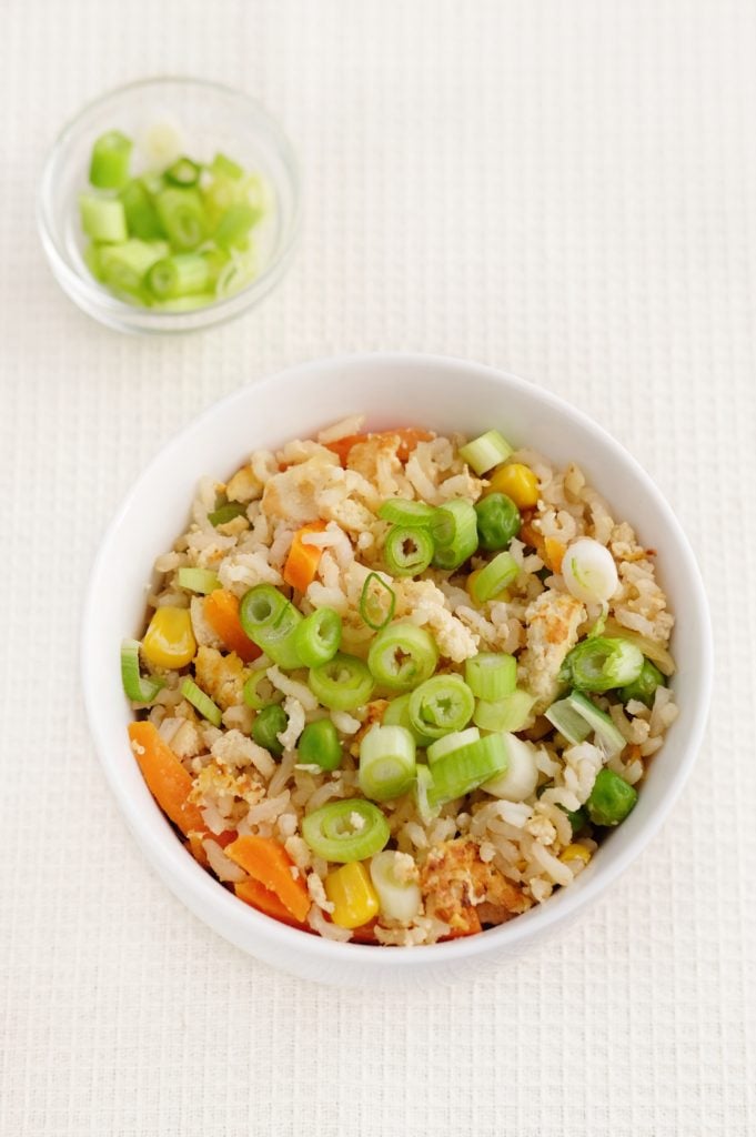 A white bowl of fried rice with diced carrots, sweetcorn, tofu and chopped spring onions, placed on a light-coloured textured surface; a small dish of spring onions is in the background.