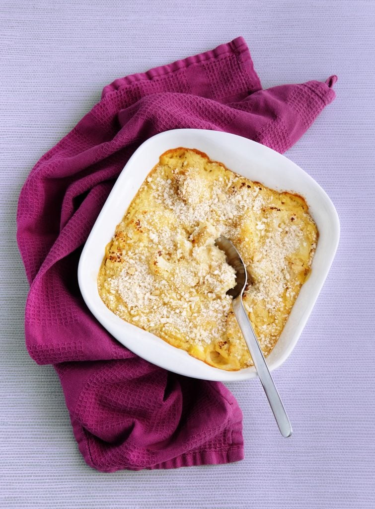 A baked casserole with a golden breadcrumb topping in a white dish, resting on a purple cloth with a spoon inside.
