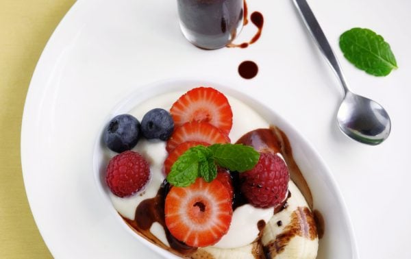 A bowl of yoghurt topped with banana slices, strawberries, blueberries, raspberries, and mint, served with chocolate sauce in a bottle and a spoon on a white plate.