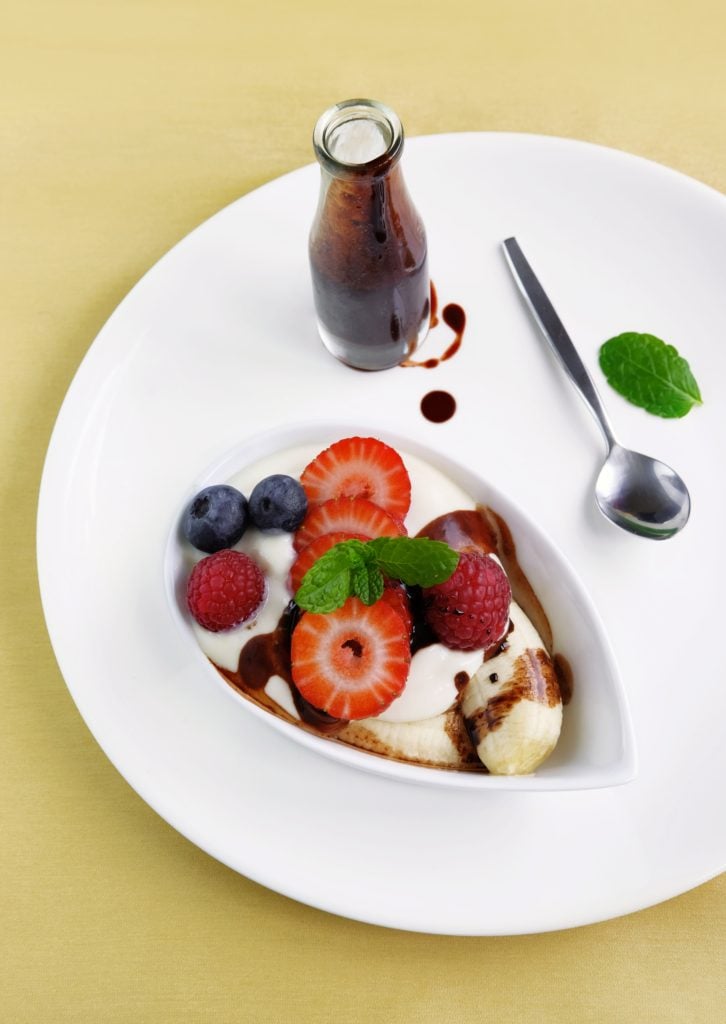 A bowl of yoghurt topped with banana slices, strawberries, blueberries, raspberries, and mint, served with chocolate sauce in a bottle and a spoon on a white plate.