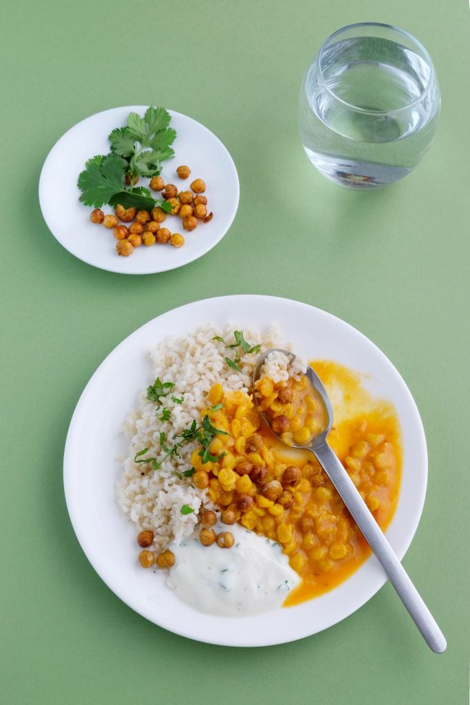 A plate with rice, chickpea curry, yoghurt sauce, and a spoon, next to a small plate of coriander and chickpeas, and a glass of water on a green surface.