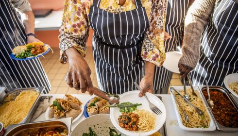 People wearing striped aprons serve food, including pasta, greens, and vegetable dishes, onto plates at a buffet-style meal.