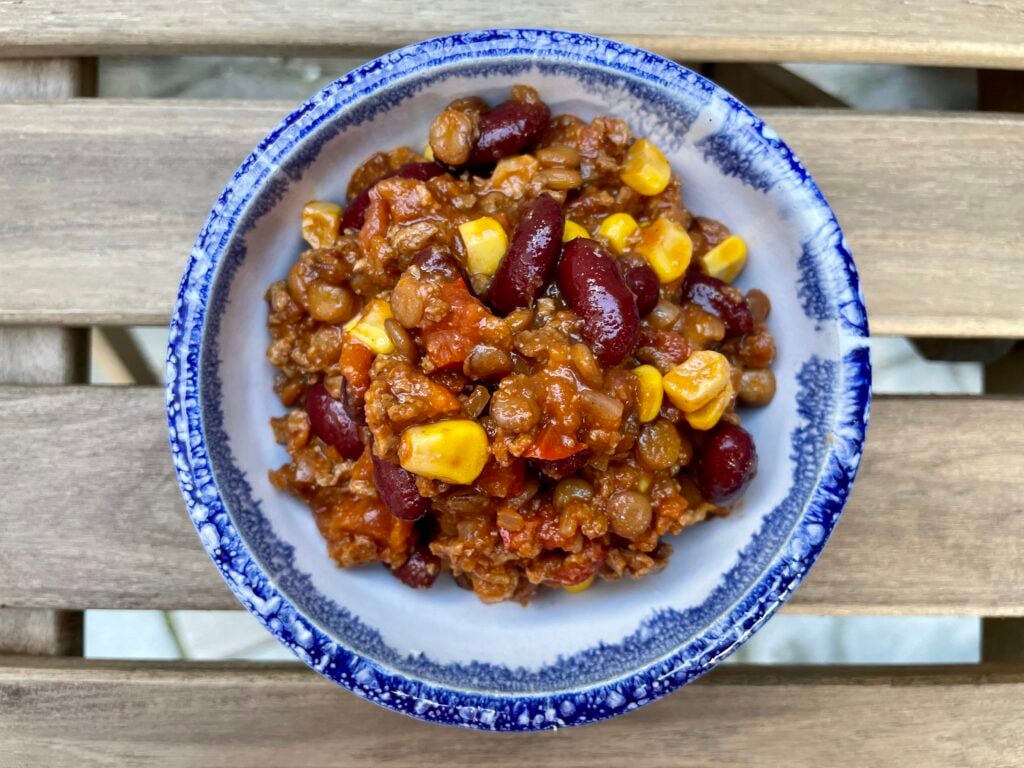 A bowl of chilli containing kidney beans, sweetcorn, minced meat, lentils, and tomatoes, served in a blue-rimmed ceramic bowl on a wooden surface.