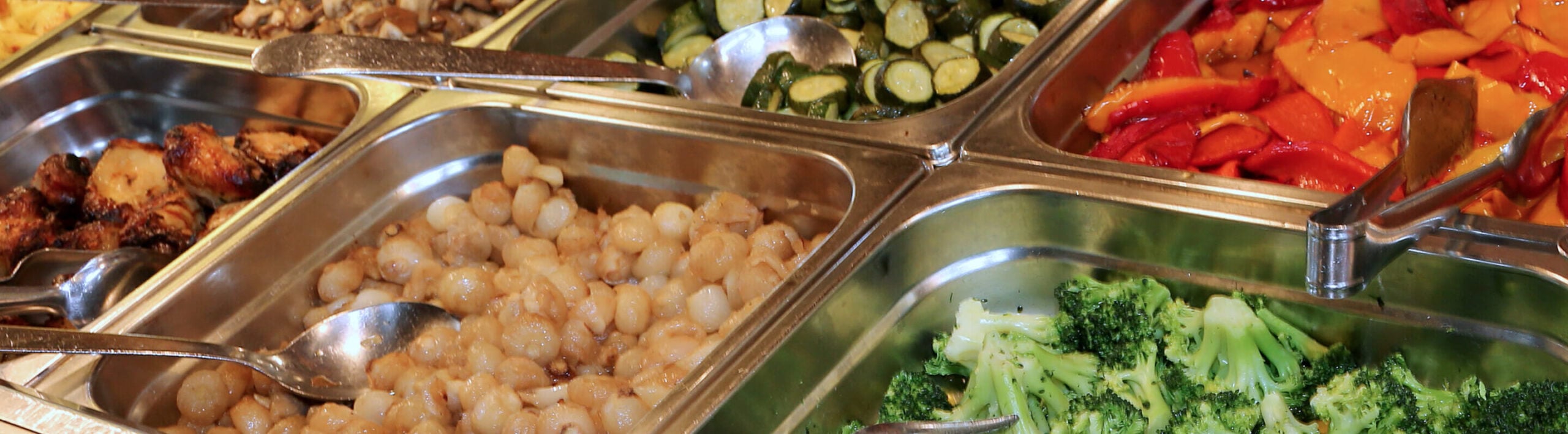 A buffet with stainless steel trays containing assorted vegetables, including broccoli, pearl onions, courgette, mushrooms, roasted peppers, and roast potatoes.