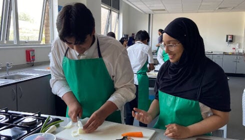 Two students wearing green aprons prepare food in a bright kitchen classroom, one chopping onions whilst the other looks on and smiles.