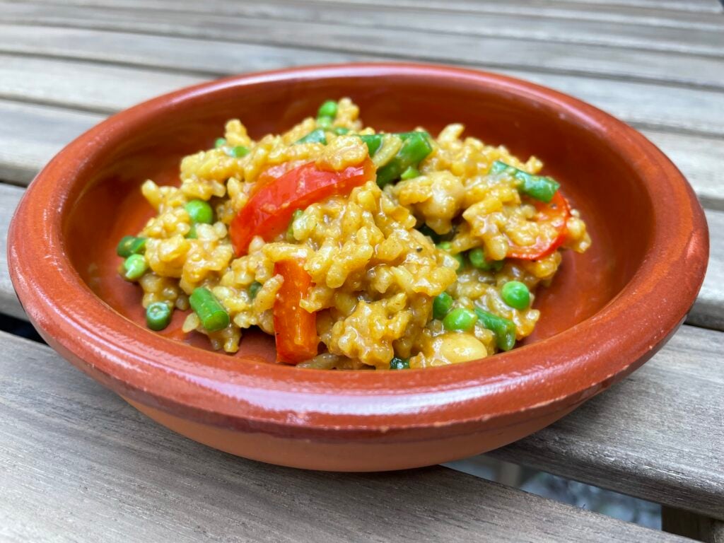 A brown ceramic bowl contains yellow rice mixed with green peas and red pepper slices, placed on a wooden surface.