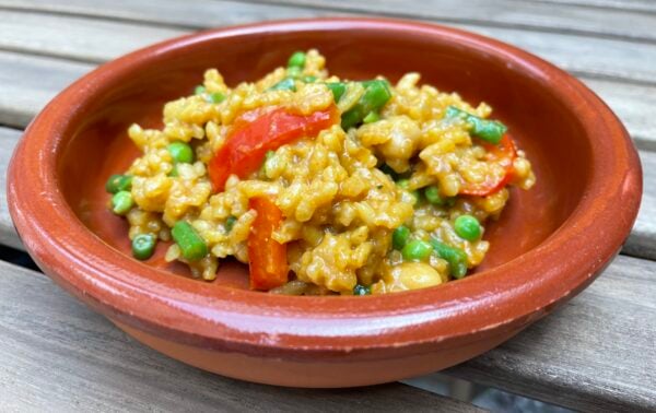 A brown ceramic bowl contains yellow rice mixed with green peas and red pepper slices, placed on a wooden surface.
