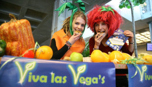 Two people dressed in playful fruit and vegetable costumes smile while eating food behind a table full of colorful produce, representing ProVeg Netherlands. A banner reading Viva las Vegas hangs in front of the table.