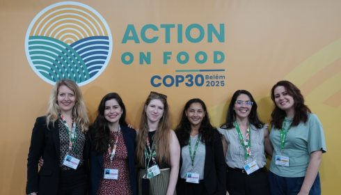 Six women stand smiling in front of a yellow backdrop with the Action on Food COP30 Belém 2025 logo, wearing conference badges and professional or casual attire.