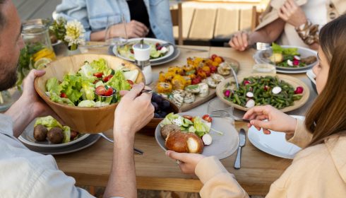 Four people sit around a wooden table sharing a meal, with plates of salad, grilled vegetables, green beans, bread, and drinks. One person serves salad from a large wooden bowl. The setting is bright and casual.