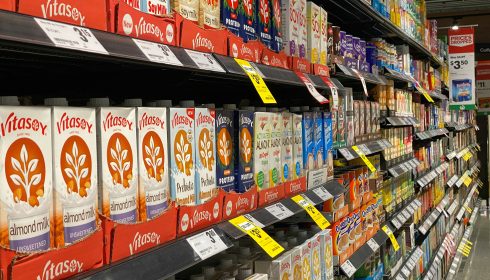 Grocery store aisle with shelves stocked with various brands and flavors of plant-based milk, including almond, soy, and oat milk in colorful cartons, with price tags visible.