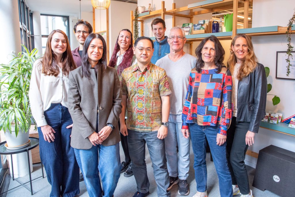 A group of nine people, both men and women, stand smiling together in a brightly lit modern office space with plants and shelves in the background.