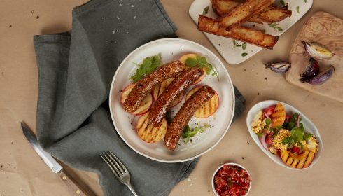 A plate with grilled sausages and sliced grilled peaches on greens, set on a gray cloth. Surrounding sides include thick potato wedges, a colorful salad, salsa, and roasted onions, with a fork and knife nearby.