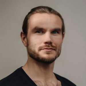 A man with light skin, brown hair tied back, a short beard, and mustache looks directly at the camera. He is wearing a dark shirt and is posed in front of a plain, light-colored background.