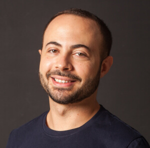 A man with short dark hair, a beard, and mustache is smiling softly. He is wearing a dark shirt and is posed against a plain dark background.