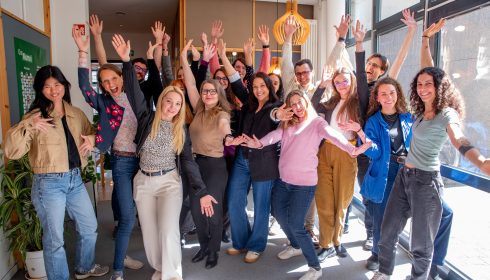 A group of people stand indoors near large windows, smiling and enthusiastically raising their arms in playful poses, reflecting the cheerful and energetic spirit inspired by the ProVeg 2026 mission and vision.