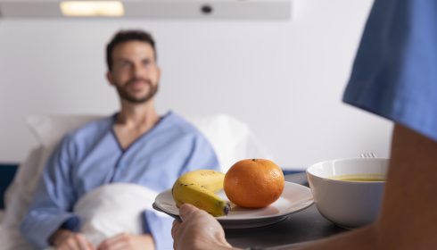 A nurse hands a plate with a banana and an orange, along with a bowl of soup, to a smiling patient sitting up in a hospital bed, wearing a blue gown. The background shows hospital equipment.