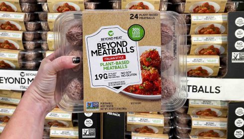 A hand holds a pack of Beyond Meat Italian Style Plant-Based Meatballs in front of a shop shelf stocked with the same product. The packaging highlights 19g protein per serving and notes that it contains no soya or gluten.