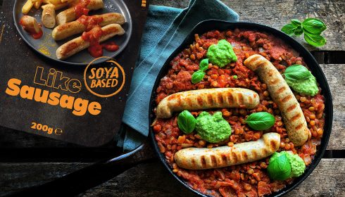 plant-based labelling vote. A frying pan with grilled vegan sausages, a chunky tomato-lentil sauce, and green pesto, next to a Like Meat Like Sausage soya-based sausage packet on a rustic wooden surface.