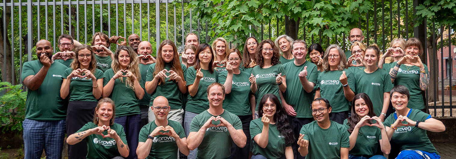 A group of people wearing matching green shirts pose outside in front of trees and a metal fence, making heart shapes with their hands and smiling at the camera.